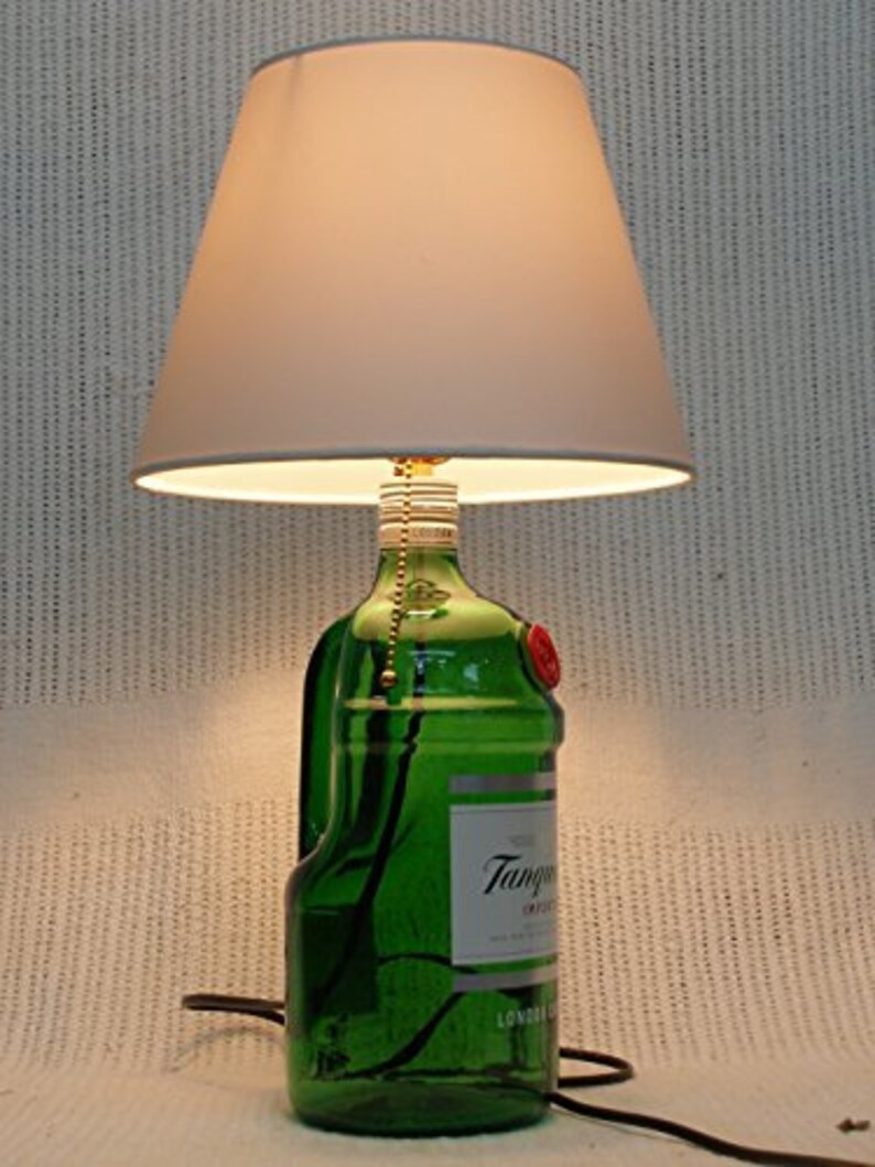 May include: A white lampshade with a green glass bottle base. The bottle is labeled "Tanqueray" and has a red wax seal. The lamp is turned on and illuminating the room.