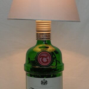 May include: A green glass bottle of Tanqueray London Dry Gin with a gold cap and a red seal. The bottle is sitting on a white surface under a white lampshade.