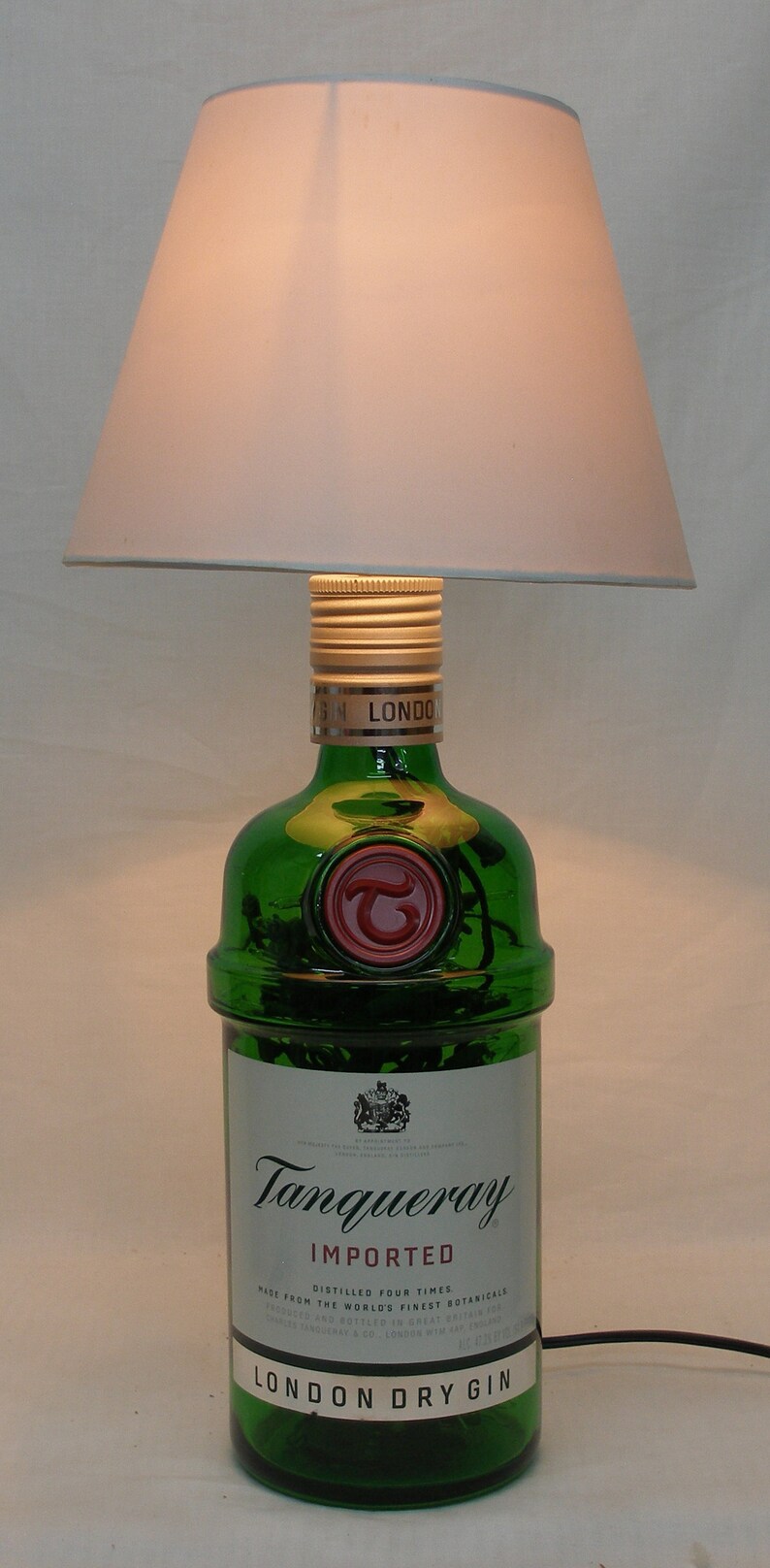 May include: A green glass bottle of Tanqueray London Dry Gin with a gold cap and a red seal. The bottle is sitting on a white surface under a white lampshade.