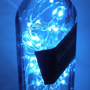 K&ouml;nnte beinhalten: Eine durchsichtige Glasflasche mit einem blauen Etikett, auf dem "Blue Label" steht, ist mit wei&szlig;en Lichterketten gef&uuml;llt. Die Flasche steht auf einer blauen Oberfl&auml;che.