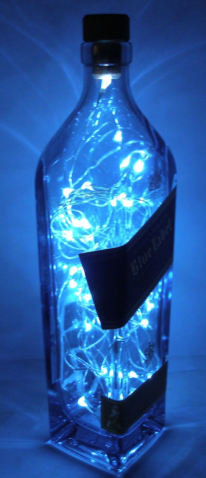 K&ouml;nnte beinhalten: Eine durchsichtige Glasflasche mit einem blauen Etikett, auf dem "Blue Label" steht, ist mit wei&szlig;en Lichterketten gef&uuml;llt. Die Flasche steht auf einer blauen Oberfl&auml;che.