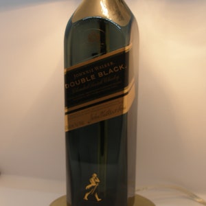 May include: A table lamp made from a repurposed Johnnie Walker Double Black Blended Scotch Whisky bottle. The lamp has a white lampshade and a gold base.