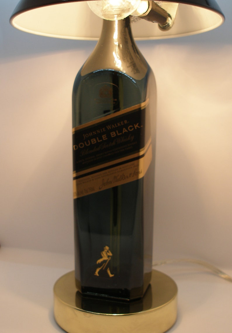 May include: A table lamp made from a repurposed Johnnie Walker Double Black Blended Scotch Whisky bottle. The lamp has a white lampshade and a gold base.
