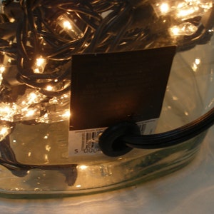 May include: A clear glass bottle with a black cord and a black label with a barcode. The bottle is filled with white Christmas lights.