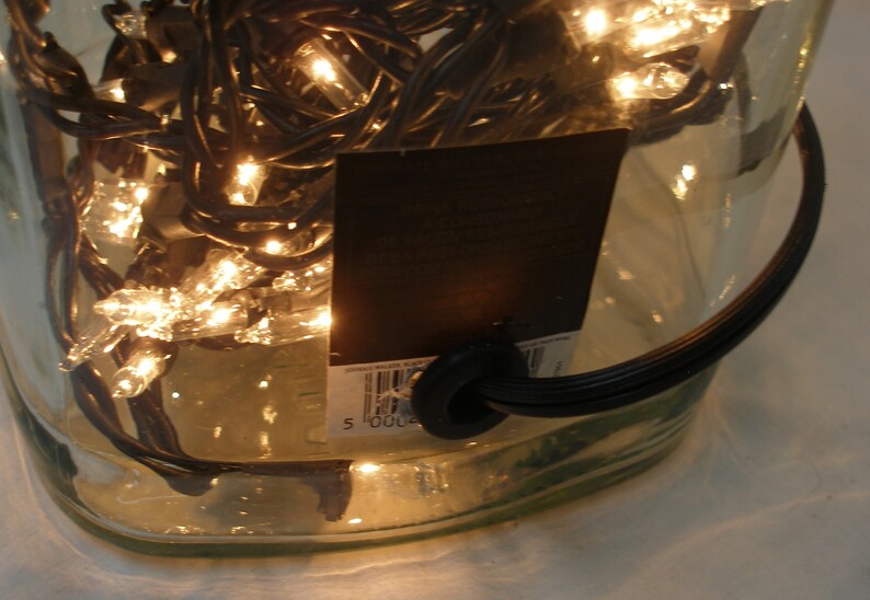 May include: A clear glass bottle with a black cord and a black label with a barcode. The bottle is filled with white Christmas lights.