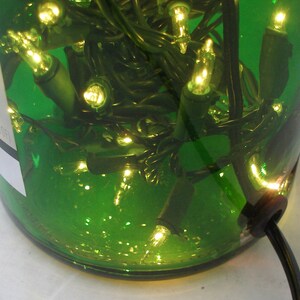 May include: A green glass bottle with a string of white Christmas lights inside. The bottle is lit up and the lights are visible through the glass. The bottle has a black cord coming out of the bottom.