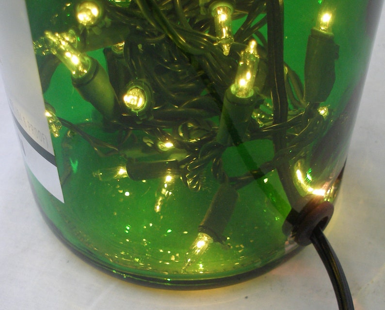 May include: A green glass bottle with a string of white Christmas lights inside. The bottle is lit up and the lights are visible through the glass. The bottle has a black cord coming out of the bottom.