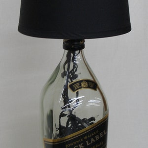 May include: A clear glass bottle with a black lampshade on top. The bottle is filled with black string lights and has a gold label that reads "Johnnie Walker Black Label Blended Scotch Whisky".