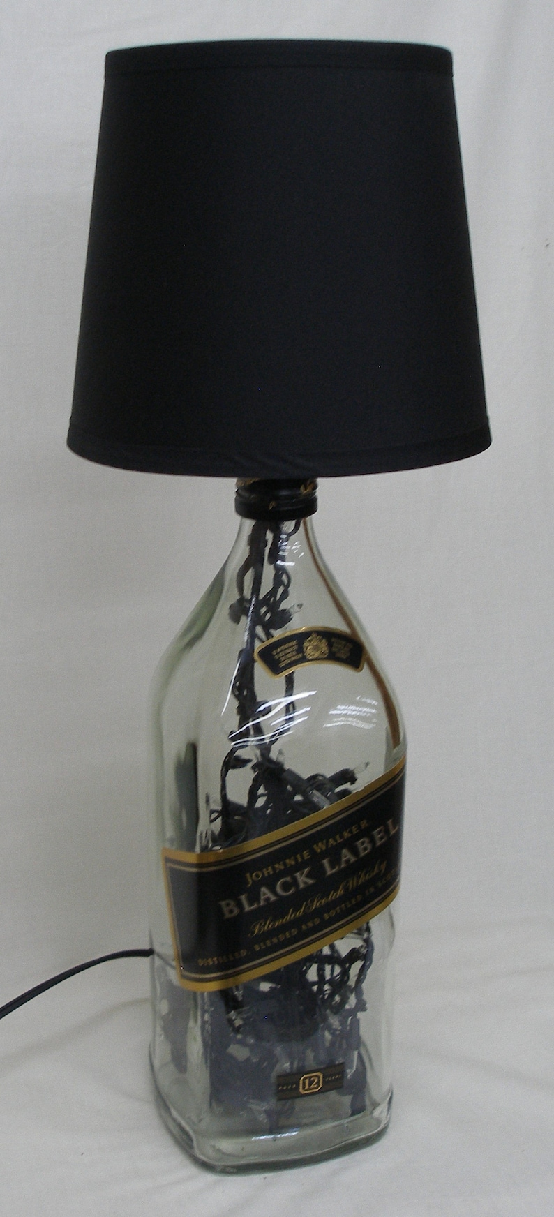 May include: A clear glass bottle with a black lampshade on top. The bottle is filled with black string lights and has a gold label that reads "Johnnie Walker Black Label Blended Scotch Whisky".
