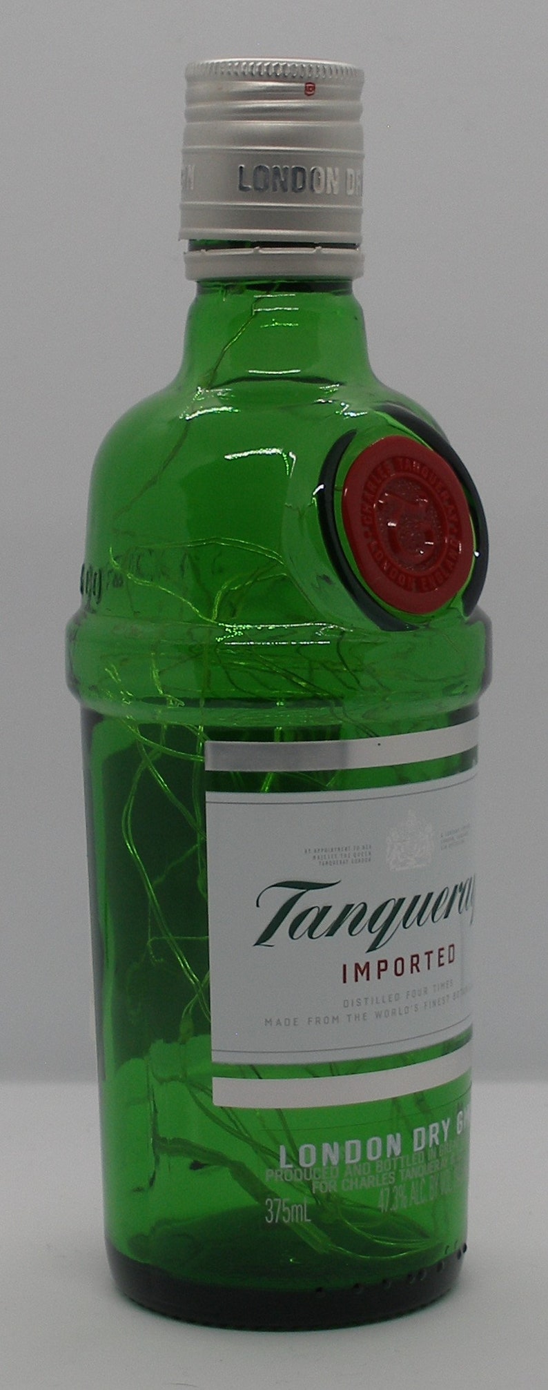 Puede incluir: Una botella de vidrio verde de Tanqueray London Dry Gin. La botella tiene una tapa plateada con el texto "LONDON DRY GIN" impreso en ella. La etiqueta es blanca con el texto "Tanqueray" en escritura verde y "IMPORTED" en negro. La botella es de 375 ml.