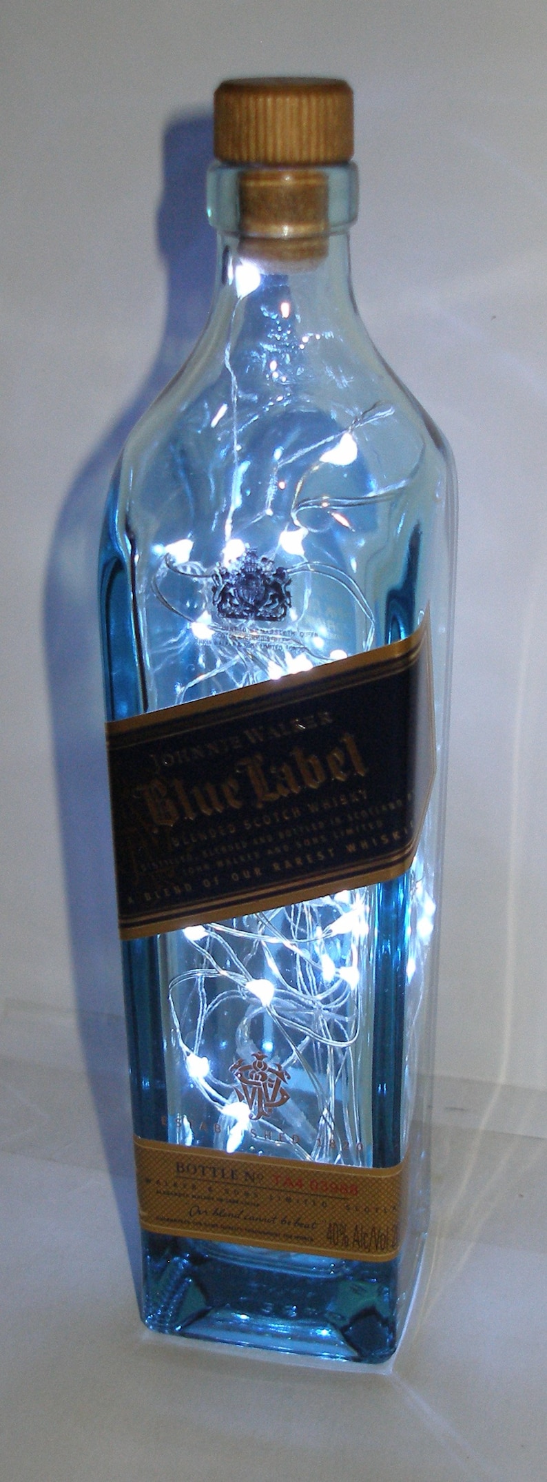 K&ouml;nnte beinhalten: Eine blaue Glasflasche mit einem goldenen Etikett, auf dem "Johnnie Walker Blue Label Blended Scotch Whisky" steht. Die Flasche ist mit wei&szlig;en Lichterketten gef&uuml;llt.