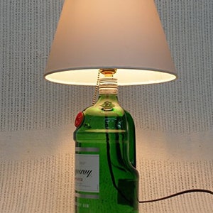 May include: A green glass bottle with a white label that reads "Tanqueray" and "London Dry Gin" is used as a lamp base. The bottle has a white lampshade.