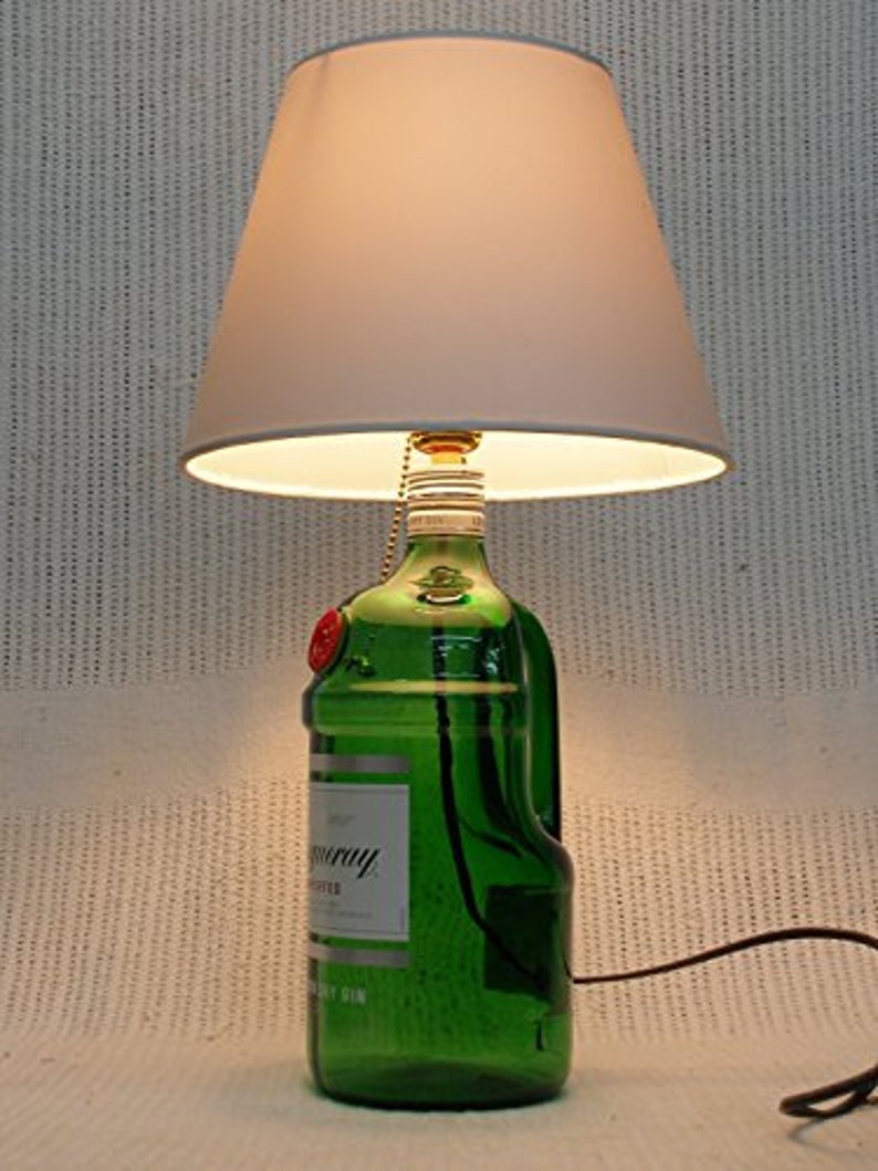 May include: A green glass bottle with a white label that reads "Tanqueray" and "London Dry Gin" is used as a lamp base. The bottle has a white lampshade.
