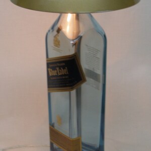 Pu&ograve; includere: Una lampada da tavolo unica realizzata con una bottiglia riciclata di Johnnie Walker Blue Label. Presenta un paralume dorato metallico e una base argentata. L'etichetta originale della bottiglia &egrave; visibile.