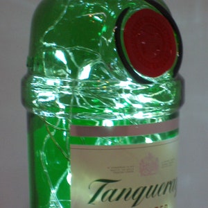 Puede incluir: Una botella de vidrio verde de Tanqueray London Dry Gin con un sello rojo y luces blancas enredadas a su alrededor. La etiqueta dice "Tanqueray Importado" y "Hecho con el mejor gin del mundo".