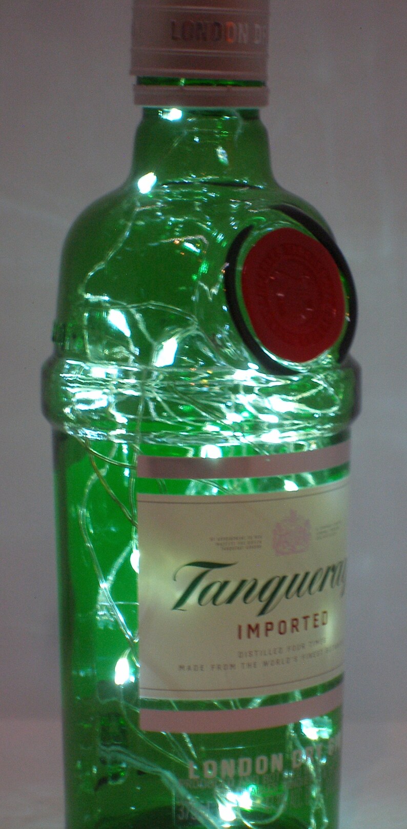 Puede incluir: Una botella de vidrio verde de Tanqueray London Dry Gin con un sello rojo y luces blancas enredadas a su alrededor. La etiqueta dice "Tanqueray Importado" y "Hecho con el mejor gin del mundo".