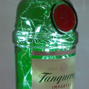 Puede incluir: Una botella de ginebra Tanqueray de vidrio verde con un sello rojo y una etiqueta blanca. La botella est&aacute; decorada con luces blancas.