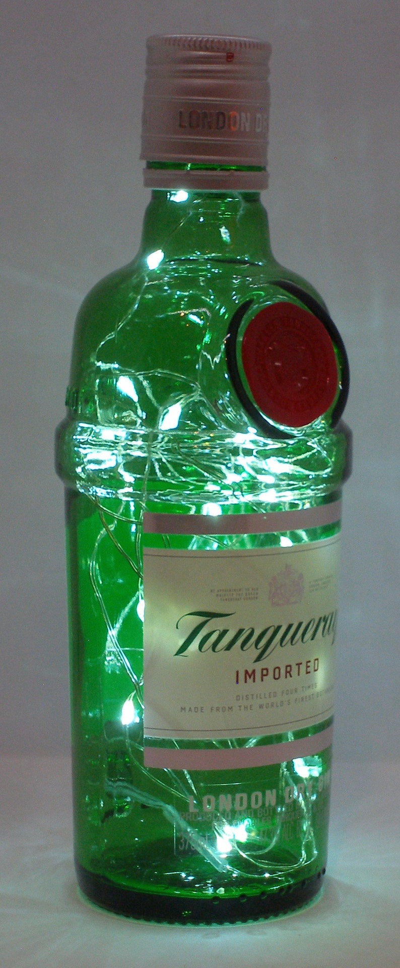 Puede incluir: Una botella de ginebra Tanqueray de vidrio verde con un sello rojo y una etiqueta blanca. La botella est&aacute; decorada con luces blancas.