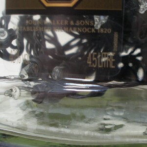 May include: A clear glass bottle with a black label that reads "AGED 12 YEARS" and "JOHN WALKER & SONS ESTABLISHED KILMARNOCK 1820". The bottle is filled with a dark liquid and has a black plastic cap.