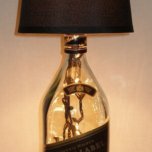 May include: A clear glass bottle with a black label that reads "Johnnie Walker Black Label Blended Scotch Whisky" is filled with white fairy lights. The bottle is topped with a black lampshade.