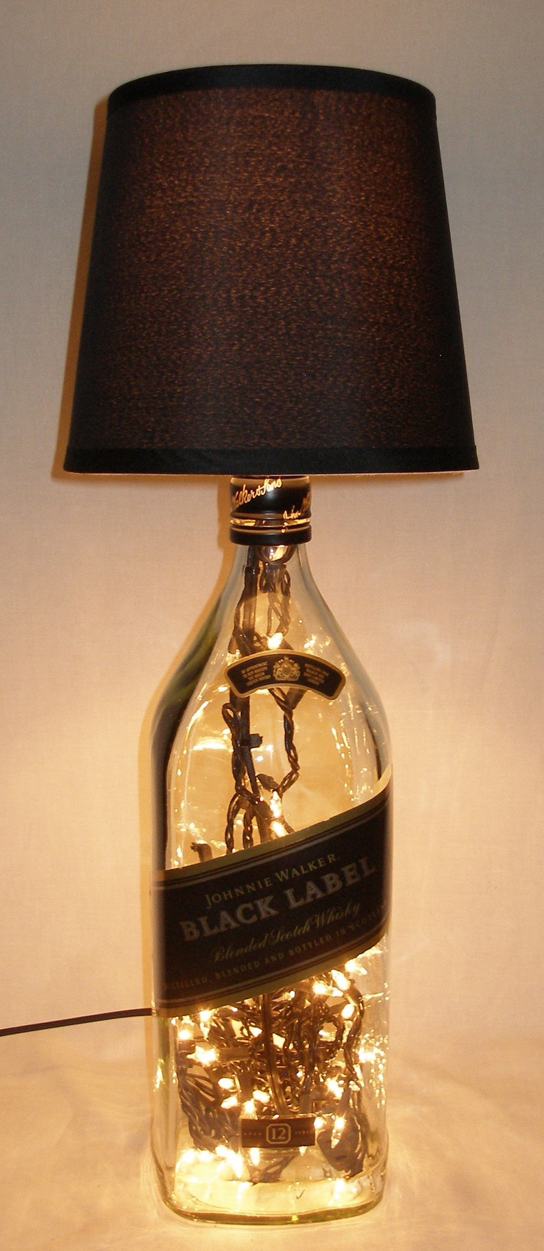 May include: A clear glass bottle with a black label that reads "Johnnie Walker Black Label Blended Scotch Whisky" is filled with white fairy lights. The bottle is topped with a black lampshade.