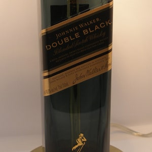 May include: A dark green glass bottle of Johnnie Walker Double Black Blended Scotch Whisky. The bottle has a gold label with the brand name and a description of the whisky. The bottle is sitting on a gold metal base.