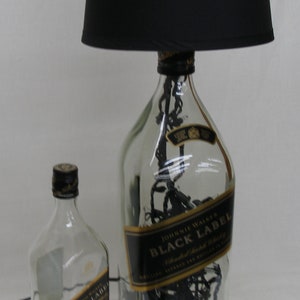 May include: A black lampshade sits atop a clear glass bottle with a black label that reads "Johnnie Walker Black Label Blended Scotch Whisky." The bottle is filled with black string lights. A smaller, clear glass bottle with a black label that reads "Johnnie Walker Black Label" sits in front of the lamp.