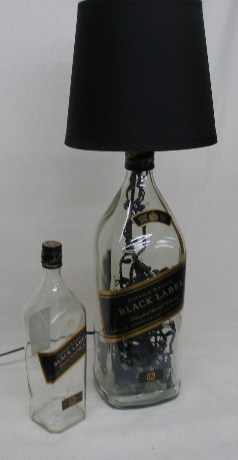 May include: A black lampshade sits atop a clear glass bottle with a black label that reads "Johnnie Walker Black Label Blended Scotch Whisky." The bottle is filled with black string lights. A smaller, clear glass bottle with a black label that reads "Johnnie Walker Black Label" sits in front of the lamp.