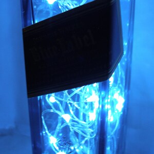 K&ouml;nnte beinhalten: Eine durchsichtige Glasflasche mit einem blauen Etikett, auf dem "Black Label" steht, ist mit blauen LED-Lichtern gef&uuml;llt.