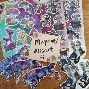 May include: A collection of stickers featuring various designs, including mushrooms, cats, and witchy themes. The stickers are arranged on a wooden surface with a note that reads "Misprint/Miscut".