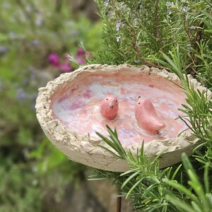 Könnte beinhalten: Eine handgefertigte Vogeltränke aus Keramik mit zwei rosa Vogelfiguren. Die Vogeltränke hat einen strukturierten, hellbeigen Rand und einen rosa-weißen Innenraum. Die Vogeltränke ist von grünem Laub umgeben und schafft eine natürliche Szene im Freien.