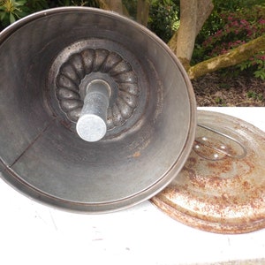 Könnte beinhalten: Eine offene, antike Gugelhupf-Form mit einem zentralen Rohr und einem geriffelten Design. Die dunkle Metallform liegt neben ihrem Deckel mit Griff. Form und Deckel weisen Gebrauchsspuren auf und haben ein rustikales Aussehen. Durchmesser ca. 25 cm.