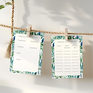 Könnte beinhalten: Vier weiße Notizblöcke mit dem Text "VERGISS MICH NICHT!" sind mit Holzklammern an einer Schnur befestigt. Die Notizblöcke haben einen dekorativen Rand aus grünen Blättern und farbigen Akzenten. Die Notizblöcke hängen vor einer weißen Wand.