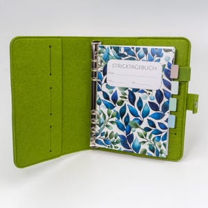 Könnte beinhalten: Offener grüner Filzordner mit silbernem Ringmechanismus. Im Inneren befindet sich ein Notizbuch mit blauem und grünem Blättermuster und dem Text "STRICKTAGEBUCH". Das Notizbuch hat farbige Registerkarten und ist auf weißem Hintergrund geöffnet.