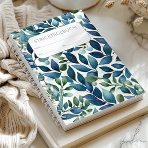 Könnte beinhalten: Ein spiralgebundenes Notizbuch mit einem Aquarellmuster aus blauen und grünen Blättern. Das weiße Etikett auf dem Einband zeigt den Text STRICKTAGEBUCH mit Zeilen für Name, Von und Bis. Das Notizbuch hat eine weiße Spiralbindung an der linken Seite.
