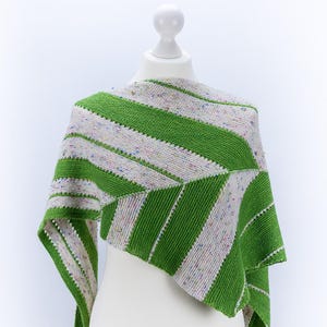 Pode incluir: Um xale de malha listrado verde e branco com um padrão geométrico. O xale está drapeado sobre um manequim branco.