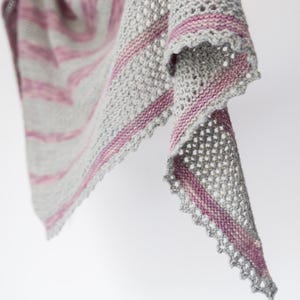 May include: A grey and pink knitted shawl with a lacy edge. The shawl is folded in half and draped over a white surface.
