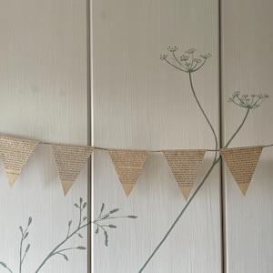 May include: A decorative banner of triangular flags made from aged book pages. The flags are strung together on a thin string. The background is a light-colored wall with a floral design in a muted green color.