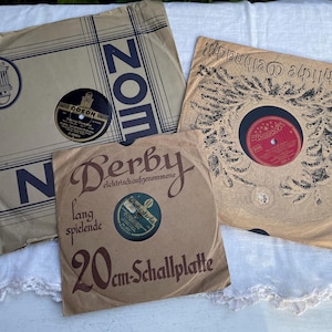 Könnte beinhalten: Drei Vintage-Vinyl-Schallplattenhüllen mit Schallplatten. Eine Hülle hat das Wort "ZOM" in Marineblau, eine andere "Derby" und "20 cm Schallplatte" in Braun, und die dritte "Fröhliche Weihnacht!" in Schwarz.