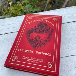 Pode incluir: Um livro de capa dura vermelho intitulado Kochbuch für drei und mehr Personen com letras douradas em relevo e uma ilustração central de pessoas a jantar. A capa apresenta bordas decorativas e texto detalhando prémios como 3 Goldene, 1 Silberne Medaille.