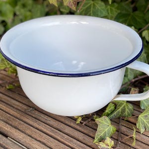Op de afbeelding: Witte emaille kom met een blauwe rand en een handvat. De kom staat op een houten oppervlak.