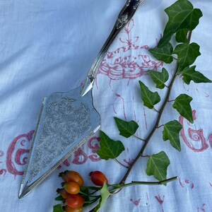 Puede incluir: Un servidor de pastel de plata con un diseño ornamentado y un mango largo. El servidor está sobre un mantel blanco con una rama de hiedra verde y bayas rojas.