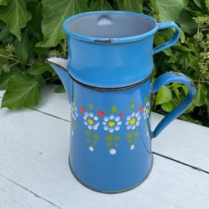 Op de afbeelding: Een vintage blauwe emaille koffiepot met bijpassende deksel. De pot is versierd met een bloemmotief van witte, gele en rode bloemen. De pot heeft een handvat en een tuit. De achtergrond is een wit houten oppervlak en groen gebladerte.