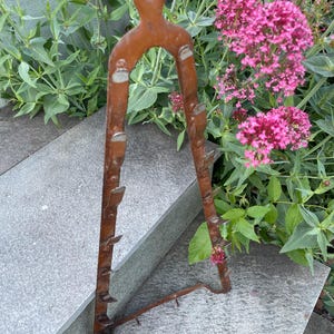 Puede incluir: Un soporte metálico triangular y oxidado que presenta una serie de pequeños ganchos curvados hacia arriba en ambos lados y un orificio en la parte superior para colgarlo. La estructura es de tiras de metal plano con acabado marrón.