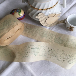 Könnte beinhalten: Eine Rolle Vintage-Papierband mit einem sich wiederholenden Muster aus Herzen und Blumen in grüner Stickerei. Eine weiße Porzellan-Teekanne, eine kleine Teetasse mit Teddybären-Design und ein dekoratives Ei sind ebenfalls sichtbar.