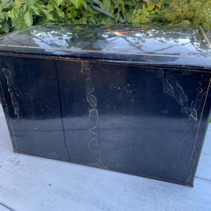 Könnte beinhalten: Eine Vintage-Metallbox in Schwarz mit leicht reflektierender Oberfläche. Die Box hat eine rechteckige Form mit flacher Oberseite und scheint gealtert zu sein, mit sichtbaren Gebrauchsspuren. Die Kanten sind mit einem goldfarbenen Akzent versehen.