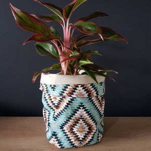 May include: A green and red houseplant in a white fabric planter with a turquoise, brown and white geometric pattern.