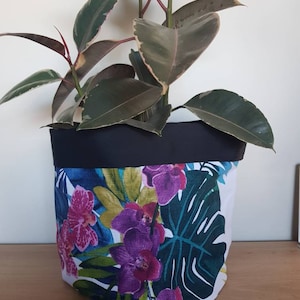 May include: A white planter with a black rim and a tropical floral print featuring pink and purple orchids and green leaves. A rubber plant with variegated leaves is growing in the planter.