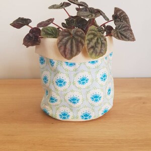 May include: A small potted plant with dark green leaves in a white fabric planter with a blue floral pattern.
