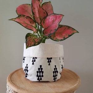 May include: A pink and green houseplant with variegated leaves in a white fabric planter with a black geometric pattern. The planter is sitting on a wooden table.